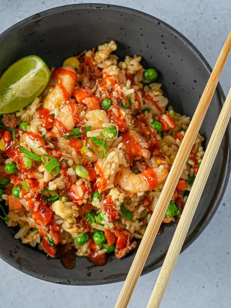 Close-up of a bowl of fried rice topped with shrimp, green onions, crispy onions, and drizzled with hot sauce, served with a lime wedge on the side