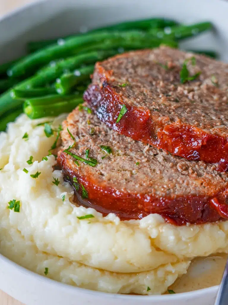 Meatloaf with a tangy tomato glaze served on a plate with mashed potatoes and green beans.