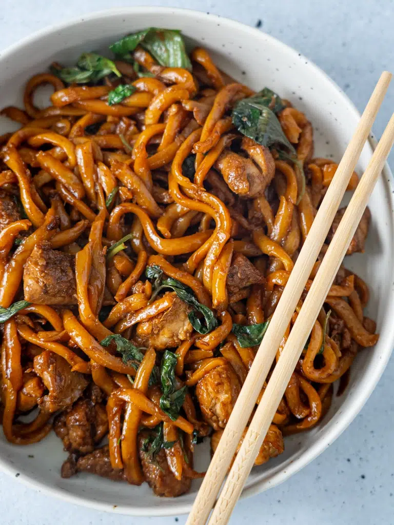Close-up of a bowl of Three-Cup Chicken Noodles with tender chicken, noodles, and a savory three-cup sauce, topped with fresh herbs and chopsticks resting on the bowl.