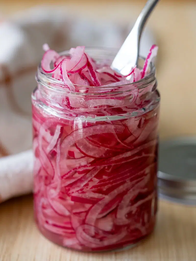 Jar of homemade pickled red onions with a fork, showcasing vibrant pink color and crisp texture.