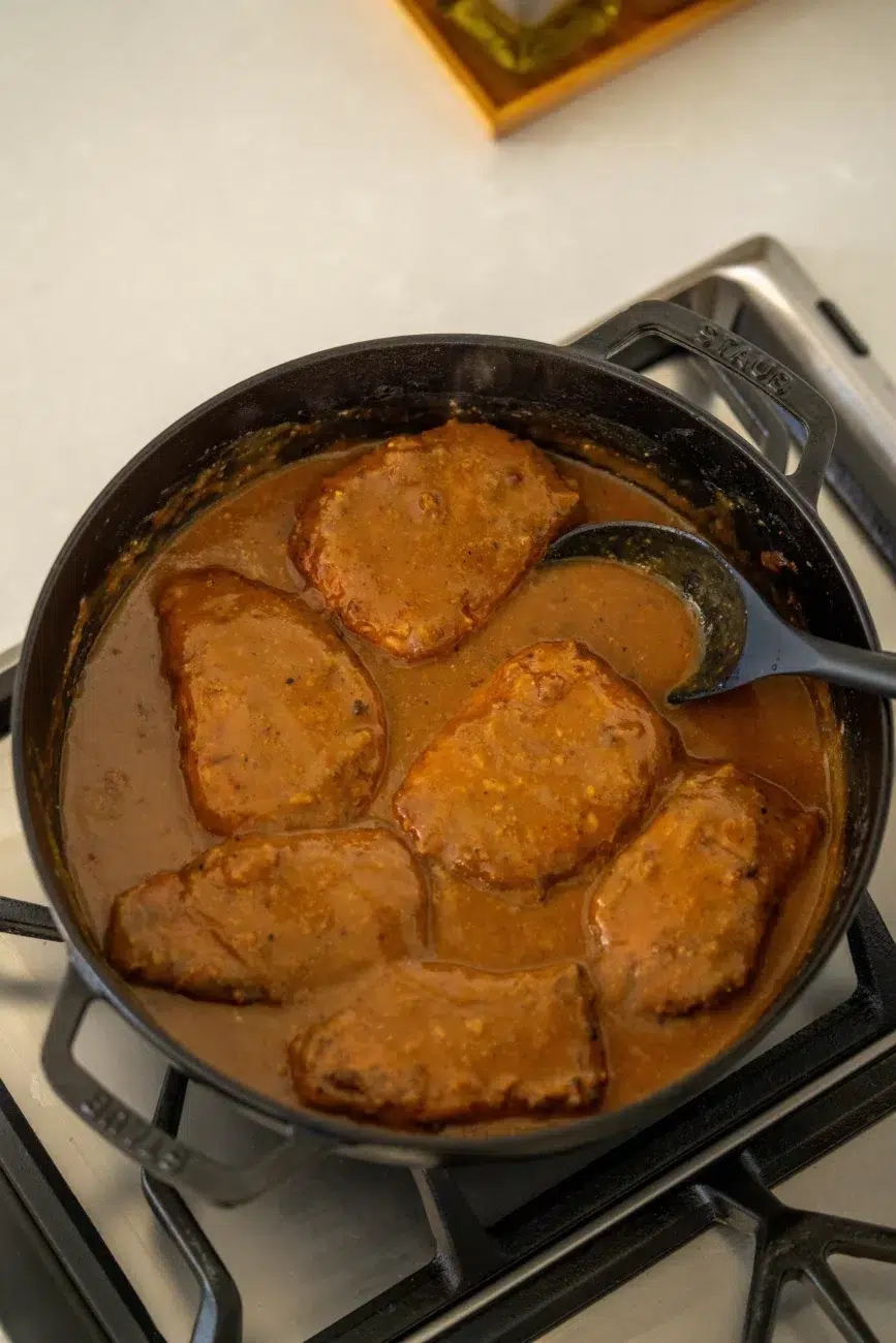 Pork chops simmering in homemade gravy inside a cast iron pan on the stovetop, with a ladle resting in the sauce