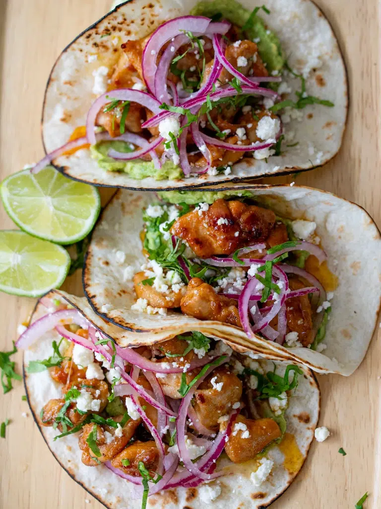 Overhead view of sticky lime honey glazed chicken tacos with pickled red onions, crumbled cheese, guacamole, and fresh herbs in toasted flour tortillas, served with lime wedges.