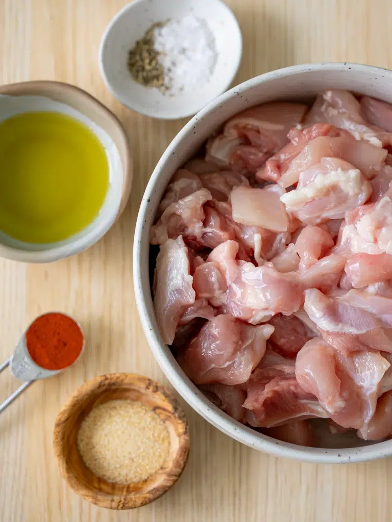 Ingredients for chicken marinade including diced raw chicken, olive oil, paprika, garlic powder, salt, and pepper arranged on a wooden surface