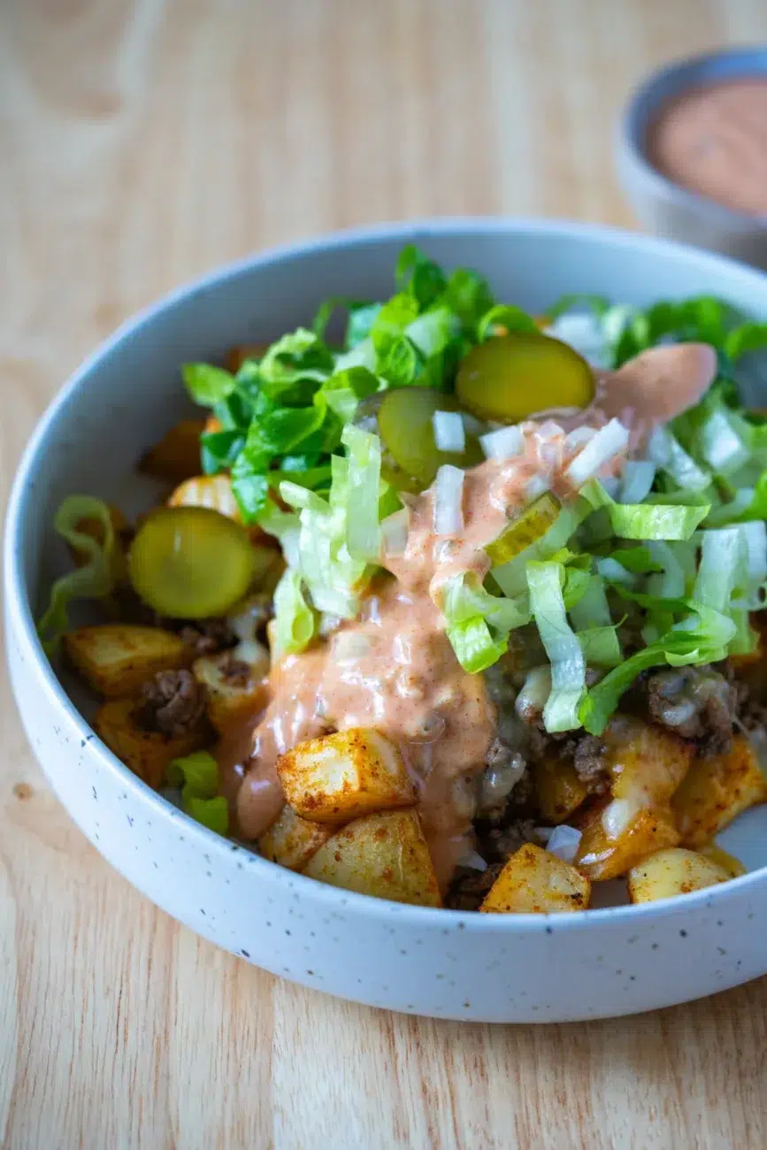Close-up of a Loaded Potato Big Mac Bowl topped with seasoned ground beef, crispy potatoes, shredded lettuce, pickles, diced onions, and creamy special sauce in a speckled ceramic bowl.