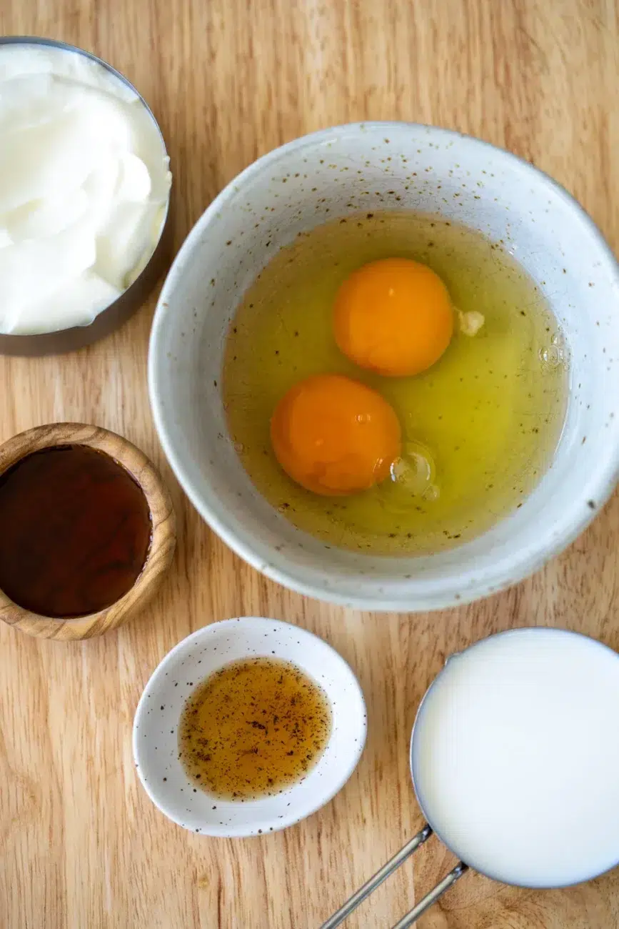 Flat lay of wet ingredients for lunchbox protein pancakes on a wooden surface, including a bowl with two cracked eggs, a measuring cup of milk, a bowl of yogurt, a small wooden bowl of vanilla extract, and a small dish with oil and black specks.