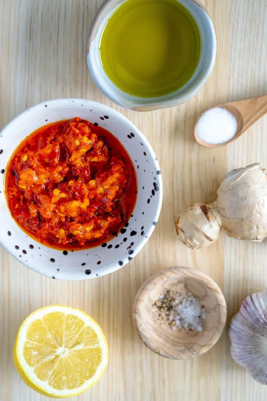 Portuguese-style chili sauce ingredients including chili oil, lemon, ginger, garlic, olive oil, salt, and pepper on a wooden surface